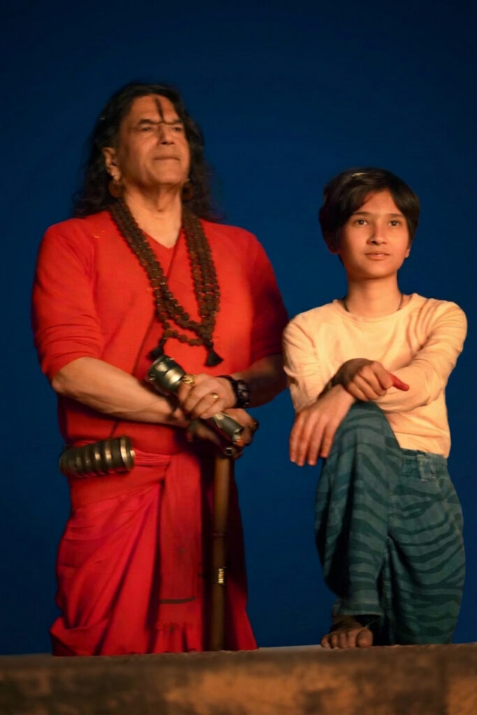 Man in a red robe with large bead necklaces and a staff standing beside a young boy on a blue backdrop.